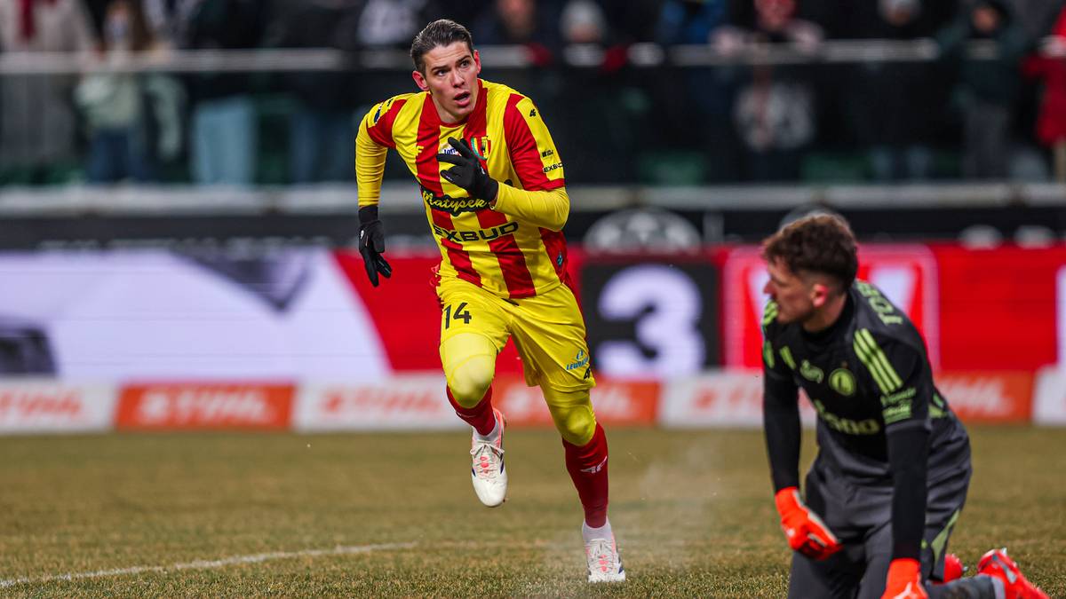 Piłkarz w ż&oacute;łto-czerwonym stroju biegnie po boisku, obok klęczy bramkarz w czarnym stroju.