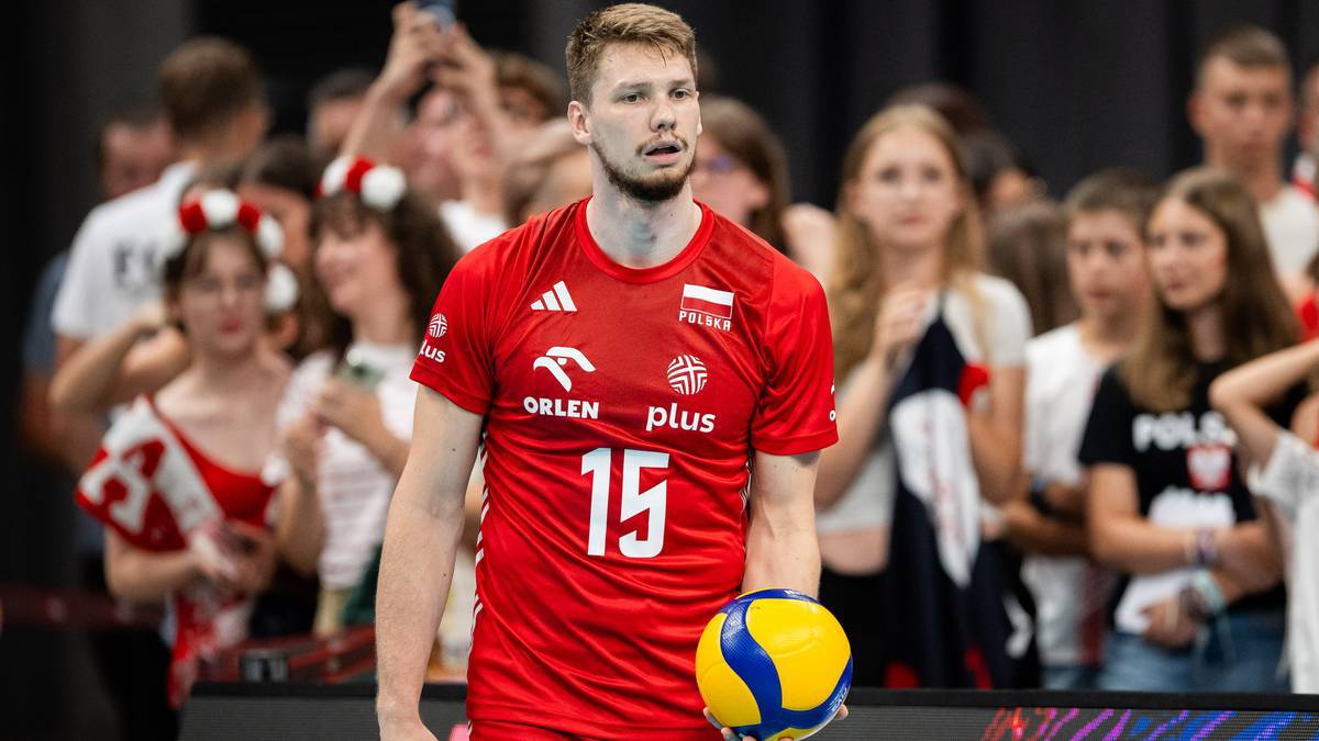 Siatkarz w czerwonej koszulce z numerem 15 i logiem Orlen Plus, trzyma piłkę do siatkówki w ręku, na tle publiczności.