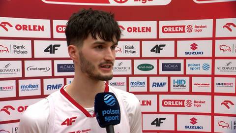 Młody mężczyzna o ciemnych włosach i z zarostem, ubrany w białą koszulkę z czerwonymi elementami, stoi przed ścianą z logo "ORLEN" i innymi sponsorami. Trzyma mikrofon "Polsat Sport" przy ustach, jakby udzielał wywiadu.