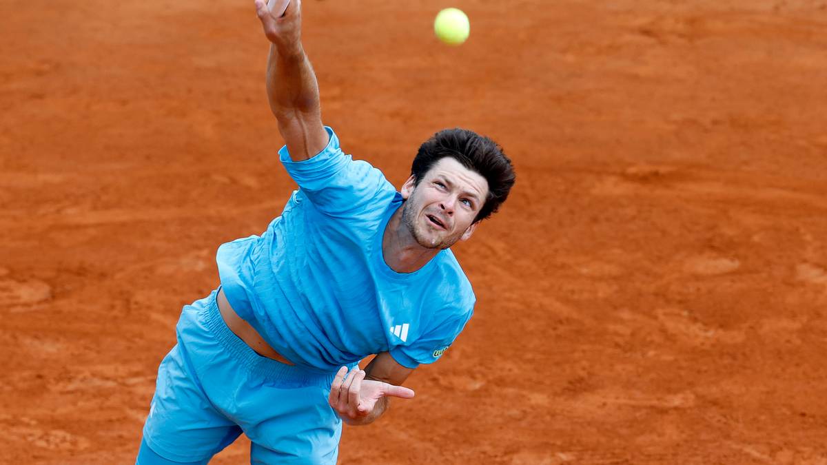 Tenisista wykonujący serwis na korcie ceglanym, w niebieskim stroju, z rakietą w g&oacute;rze i piłką w powietrzu.