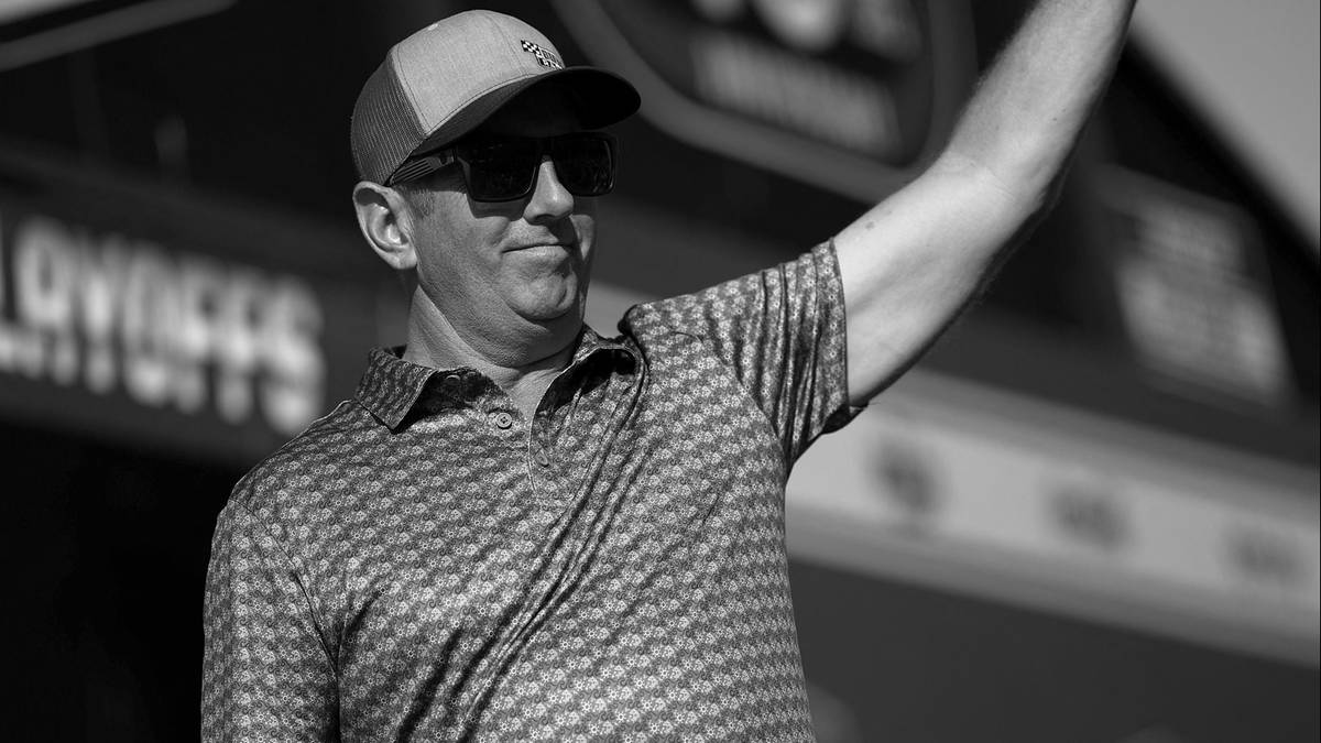 Mężczyzna w czapce i okularach przeciwsłonecznych macha ręką na tle sceny.