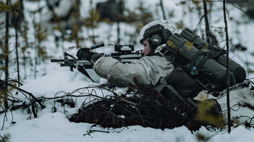 Żołnierz w zimowym kamuflażu leży w śniegu, celując z broni długiej z zamontowanym celownikiem optycznym.