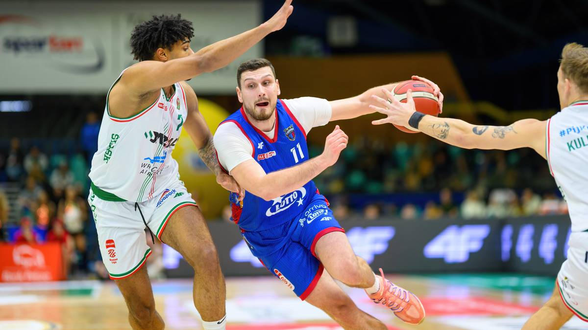 Gracz w niebieskim stroju z numerem 11 próbuje rzucić piłkę do kosza, podczas gdy dwaj przeciwnicy próbują go zablokować.