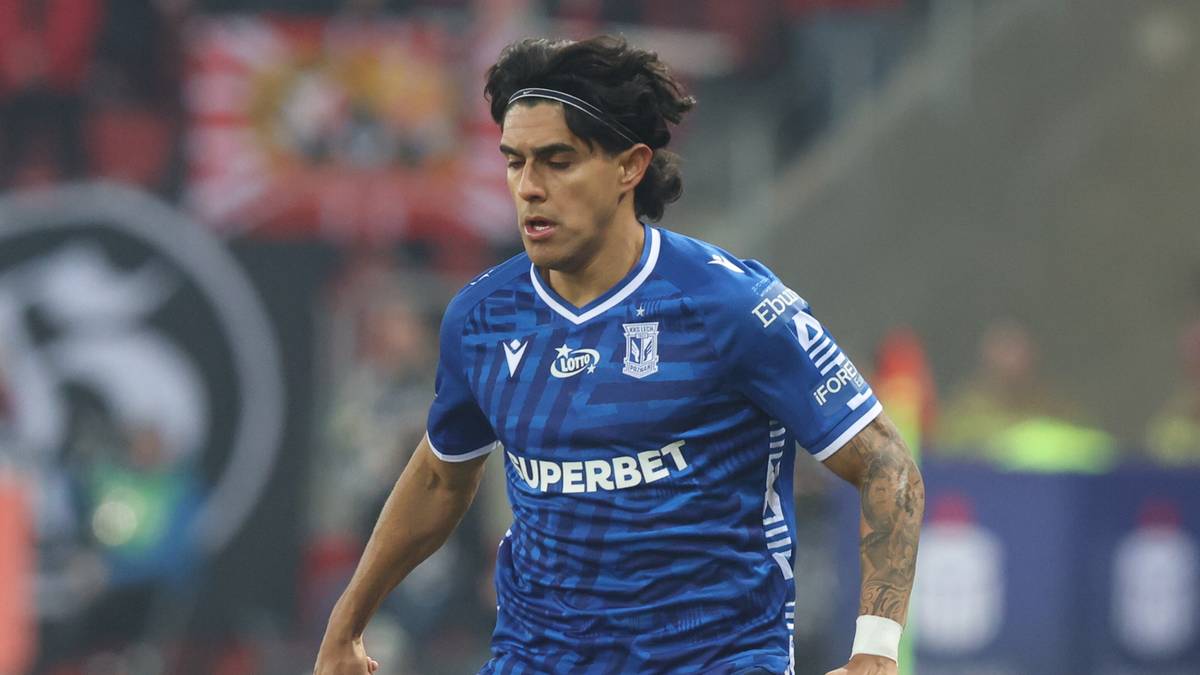 Piłkarz w niebieskim stroju z numerem 77 na plecach prowadzi piłkę na boisku.