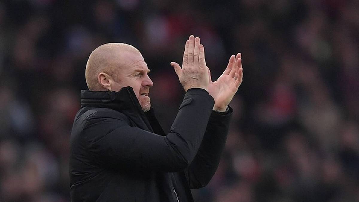 Trener piłkarski z łysą głową w czarnej kurtce z kapturem, klaszcze w dłonie. W tle widać rozmytych kibiców na stadionie.