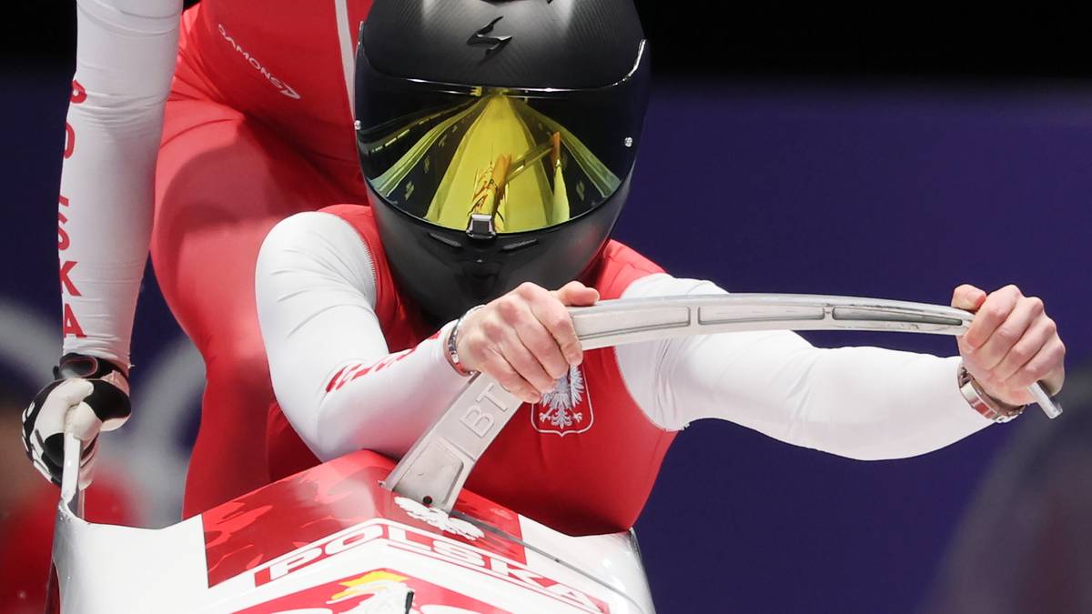 Zawodnik bobslej&oacute;w w kasku i biało-czerwonym stroju trzyma się uchwytu w pojeździe.