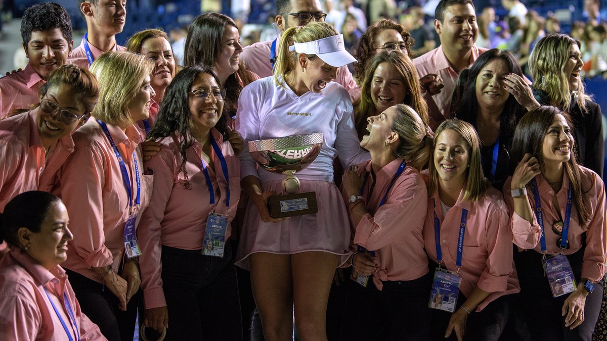 Donna Vekic w białej koszulce polo i różowej spódniczce, trzymająca puchar i śmiejąca się, otoczona przez grupę kobiet w różowych koszulkach.
