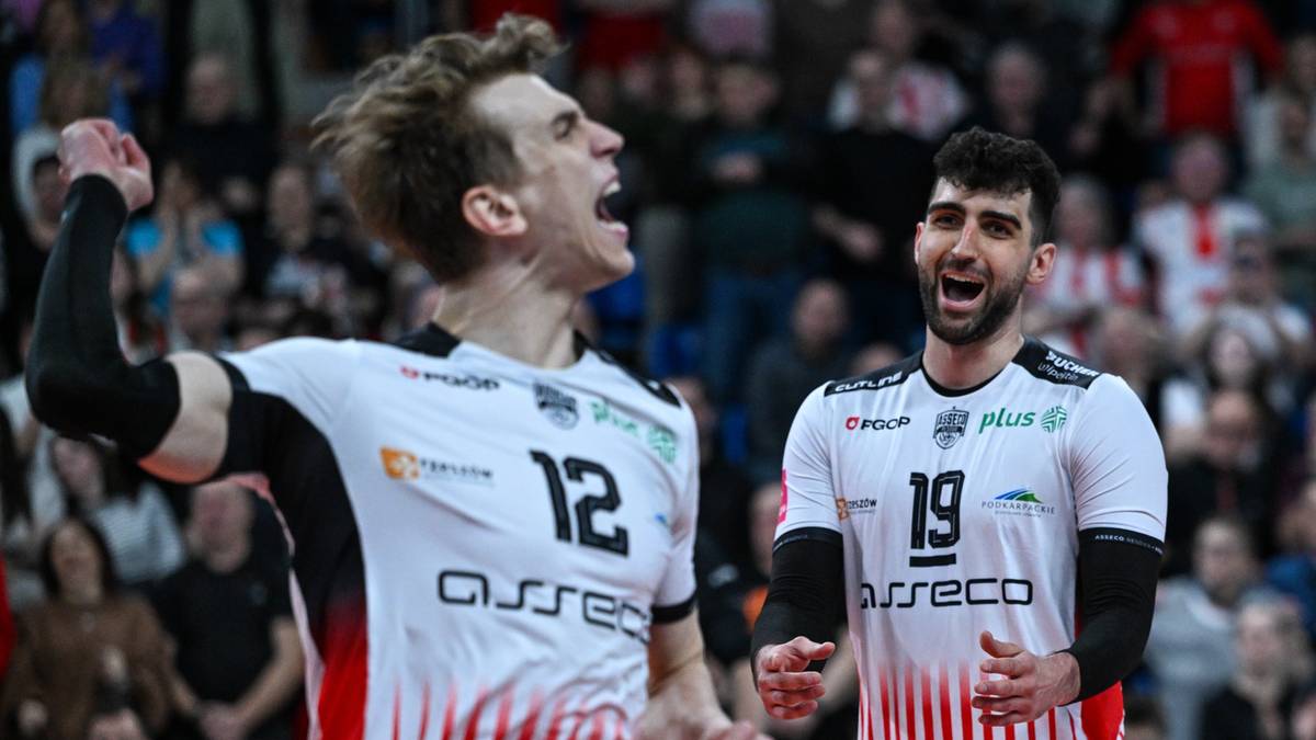 Dwóch zawodników siatkówki w białych koszulkach z czerwonymi pasami, świętujących na boisku. Jeden z nich podnosi zaciśniętą pięść, drugi krzyczy z otwartymi ustami.