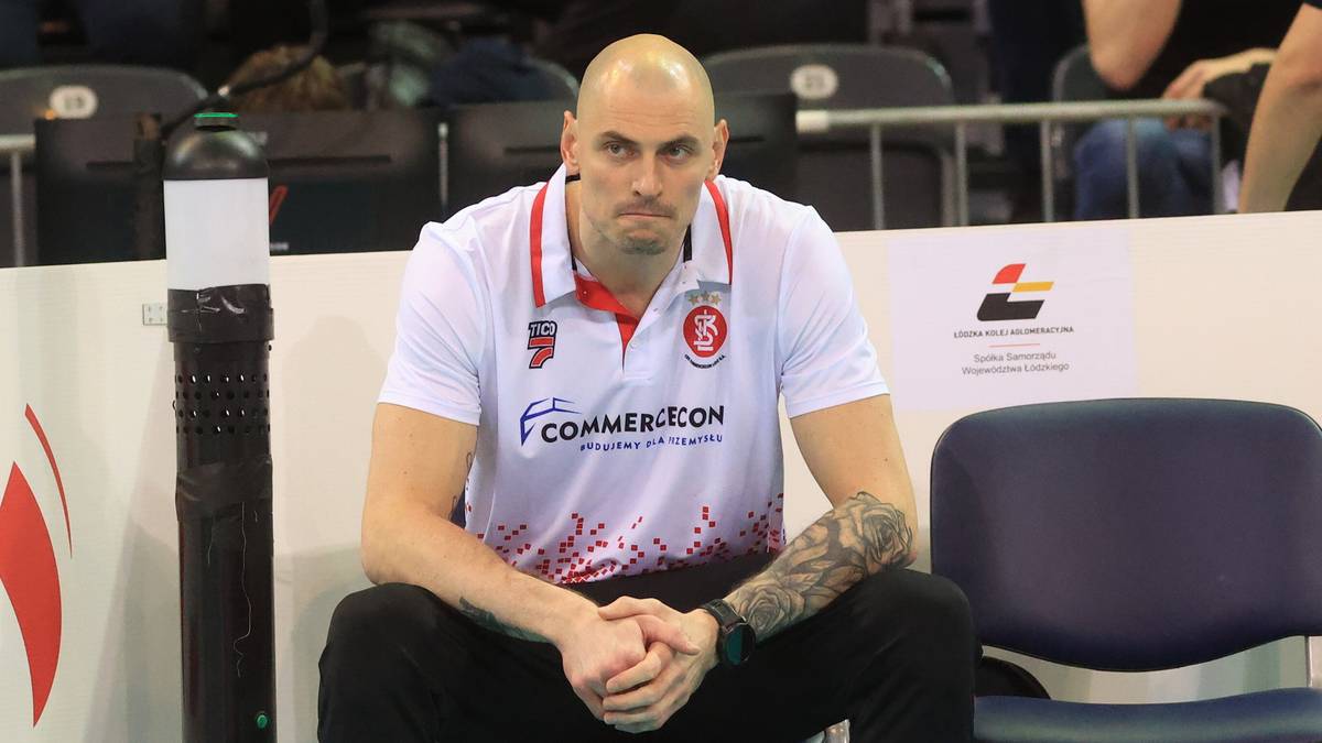 Siatkarz w białej koszulce z czerwonymi elementami siedzi na krześle w hali sportowej.