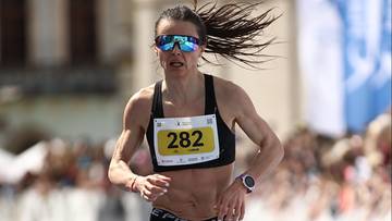 Biegaczka w ciemnej koszulce sportowej i spodenkach, z numerem startowym 282, biegnie na zawodach na zewnątrz, z włosami rozwianymi na wietrze.