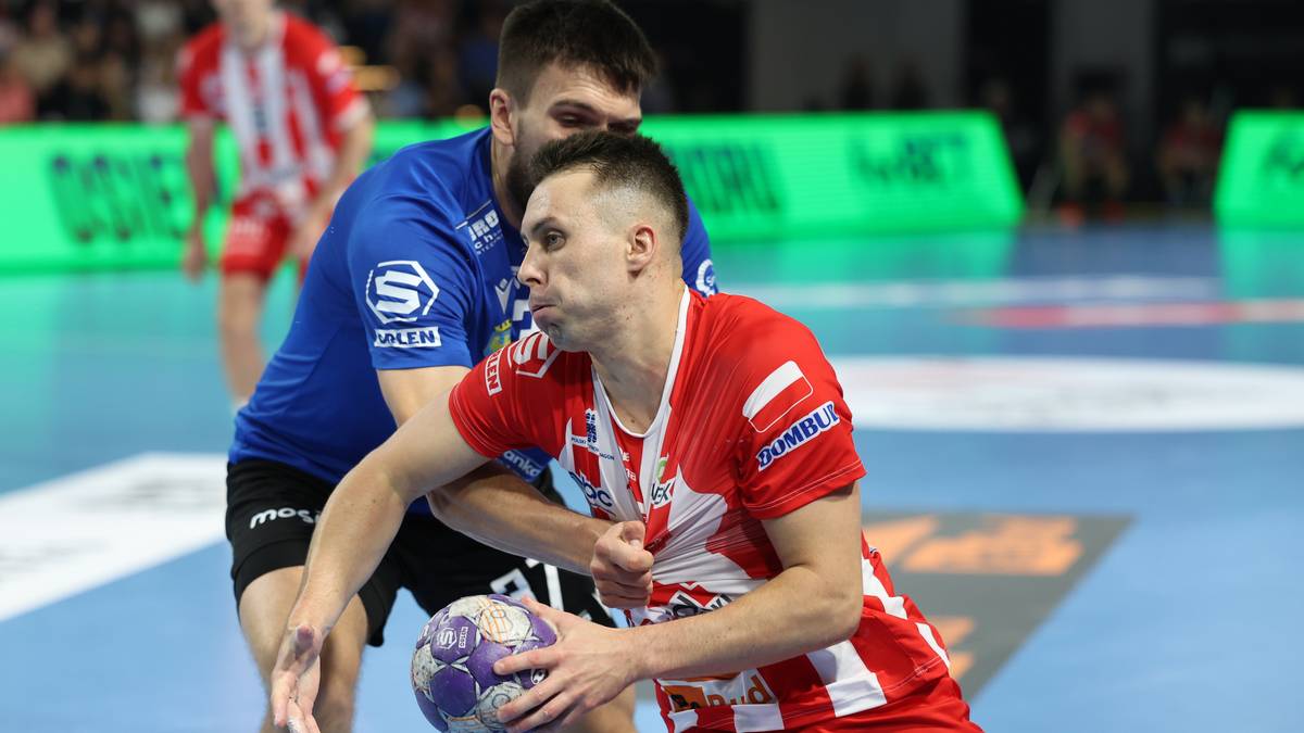 Dwóch zawodników piłki ręcznej w akcji. Jeden w czerwonej koszulce z białymi pasami próbuje rzucić piłkę, podczas gdy drugi w niebieskiej koszulce próbuje go powstrzymać.