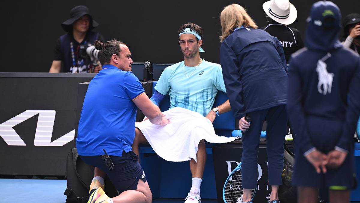 Tenisista Lorenzo Musetti siedzi z ręcznikiem na kolanach, podczas gdy fizjoterapeuta pochyla się nad jego prawą pachwiną.
