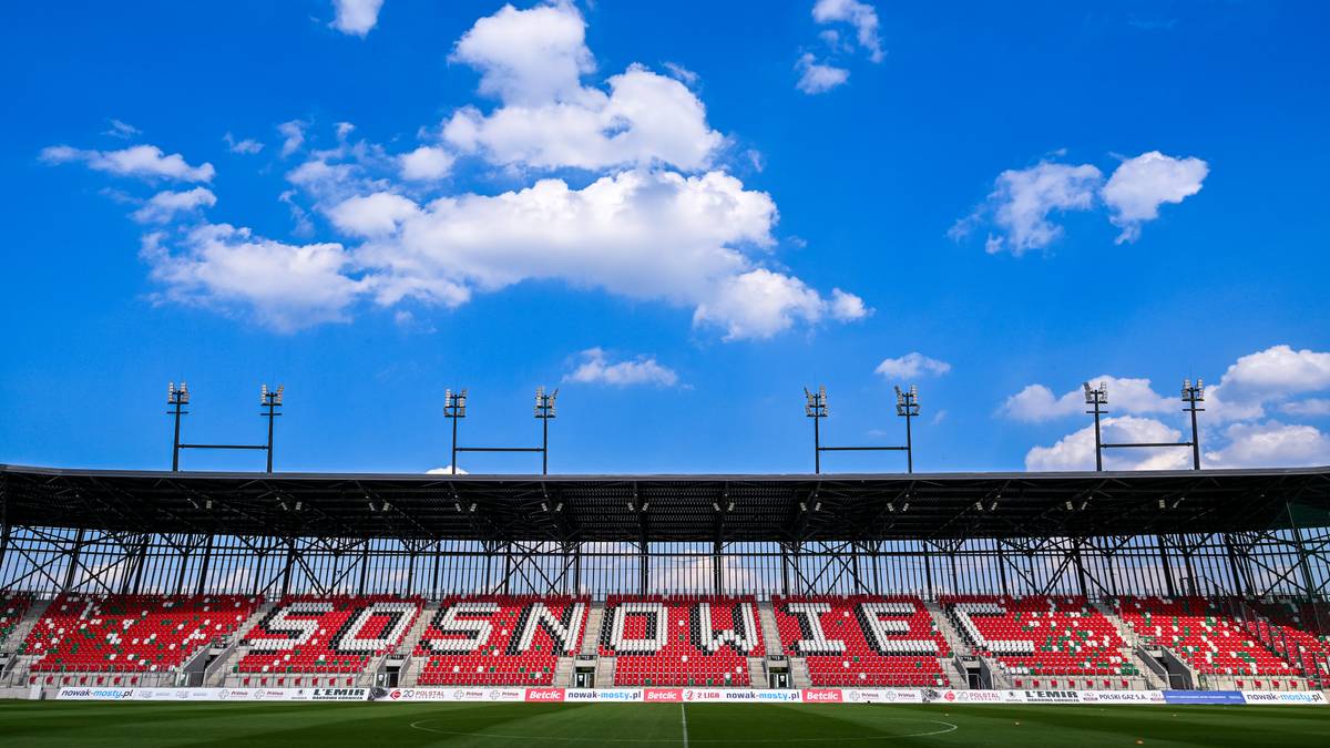 Widok na trybuny stadionu piłkarskiego z napisem SĄSIEDZI na czerwonych siedzeniach. Powyżej znajduje się zadaszenie stadionu, a w tle błękitne niebo z białymi chmurami.