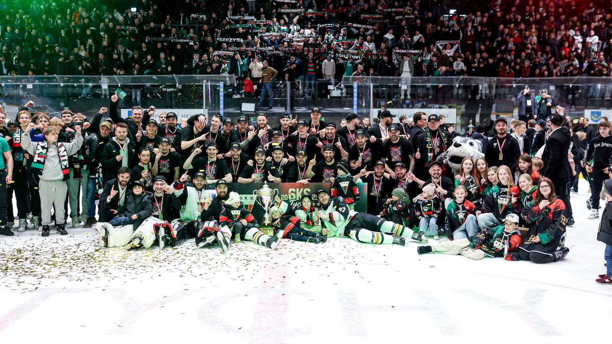 Drużyna hokejowa w komplecie, w strojach sportowych, pozuje do zdjęcia na lodowisku, w tle kibice na trybunach.