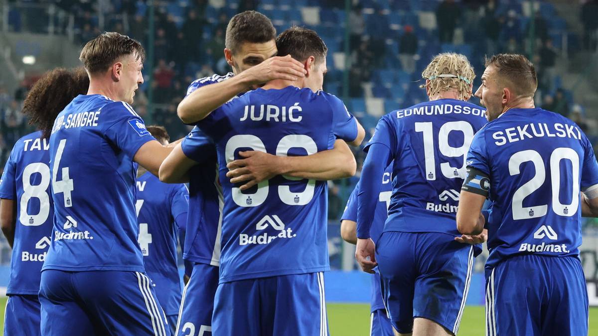 Piłkarze w niebieskich strojach świętują zwycięstwo na boisku, obejmując się i ciesząc z sukcesu.