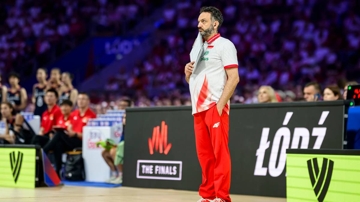 Siatkarz Stefano Lavarini w białej koszulce z czerwonymi elementami i czerwonych spodniach stoi na boisku siatkarskim, trzymając w ręku folder. W tle widać trybuny z kibicami.