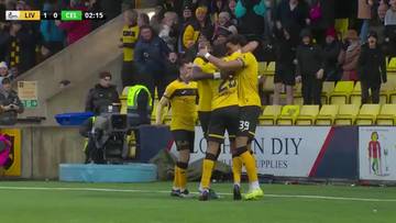 Czterech piłkarzy w ż&oacute;łtych strojach z czarnymi detalami, z numerami 39 i 35 widocznymi na koszulkach, celebruje gola, przytulając się do siebie na boisku piłkarskim. Na pierwszym planie widać ich nogi i fragmenty stroj&oacute;w, a w tle rozmyte postacie kibic&oacute;w na trybunach oraz banery.