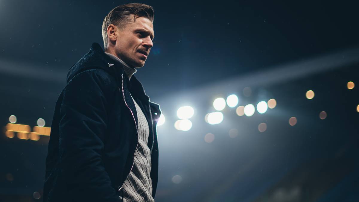 Mężczyzna w ciemnej kurtce i szarym swetrze stoi na stadionie, patrzy w bok, w tle nieostre światła.