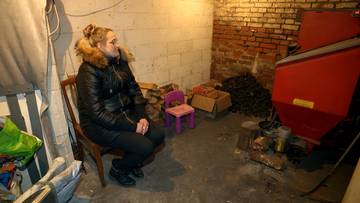 Kobieta siedzi w zimnym pomieszczeniu z piecem i opałami.