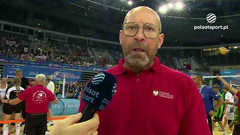 Minister Rutnicki w okularach i czerwonej koszulce, zwraca się do kamery. Na pierwszym planie widać mikrofon Polsat Sport. W tle rozmyta hala sportowa z widzami i zawodnikami.