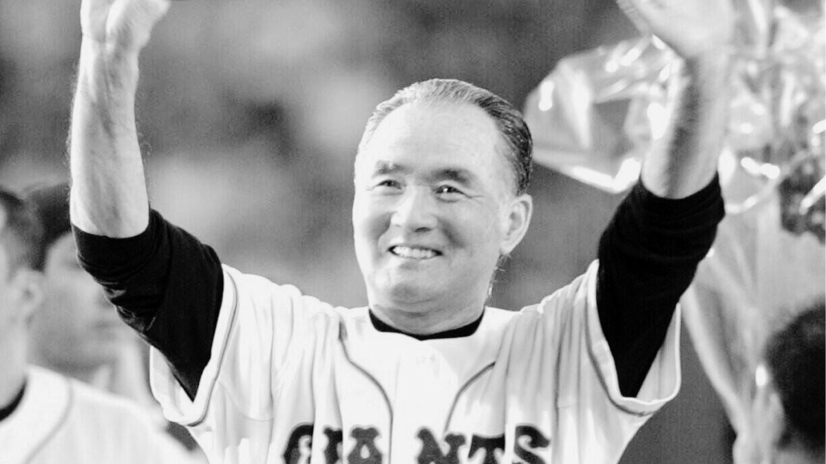 Zdjęcie czarno-białe. Mężczyzna w średnim wieku, w stroju drużyny baseballowej Giants, uśmiecha się i macha rękami na powitanie. Jego twarz jest oświetlona, a za nim widać sylwetki innych osób.