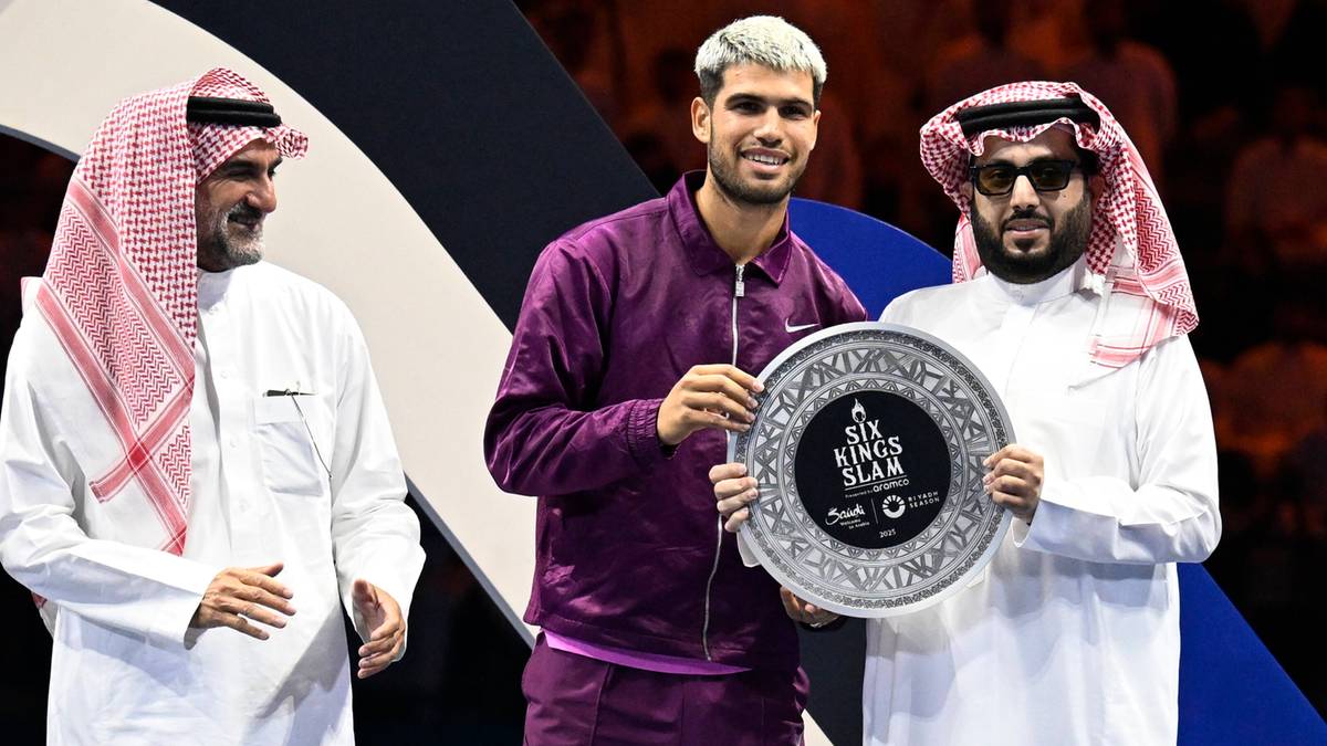 Trzech mężczyzn, z których dwóch ubranych jest w tradycyjne arabskie szaty z charakterystycznymi nakryciami głowy, wręcza okrągłą, ozdobną tarczę mężczyźnie w środku, ubranemu w ciemnofioletowy strój sportowy. Wszyscy uśmiechają się.