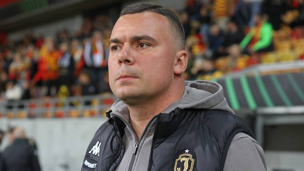 Portret mężczyzny w stroju sportowym, prawdopodobnie trenera piłkarskiego, patrzącego przed siebie z powagą. W tle widać trybuny stadionu.