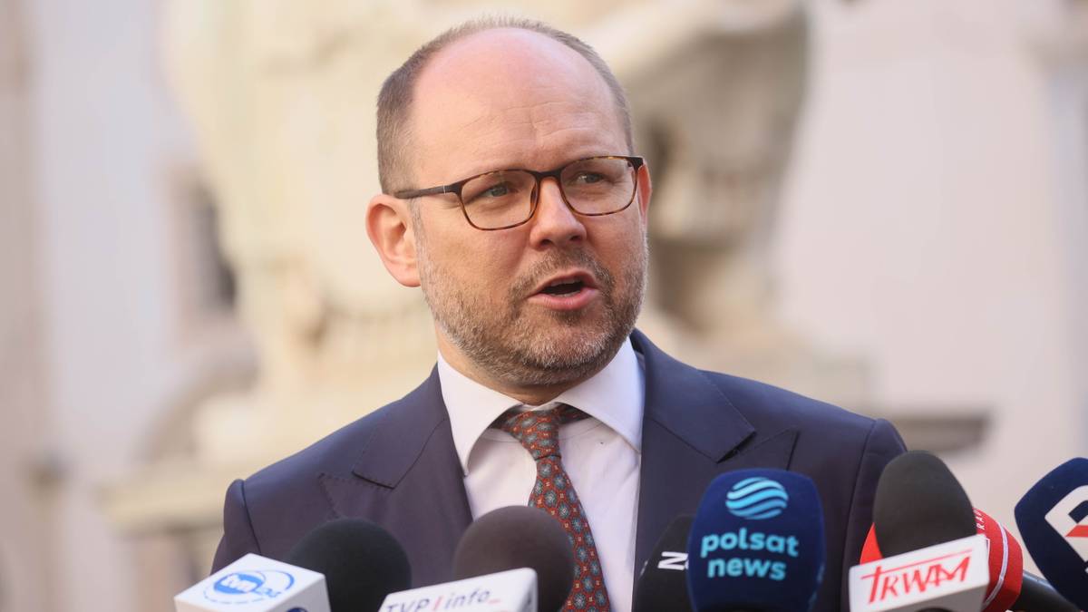 Mężczyzna w garniturze i okularach przemawia do mikrofon&oacute;w.