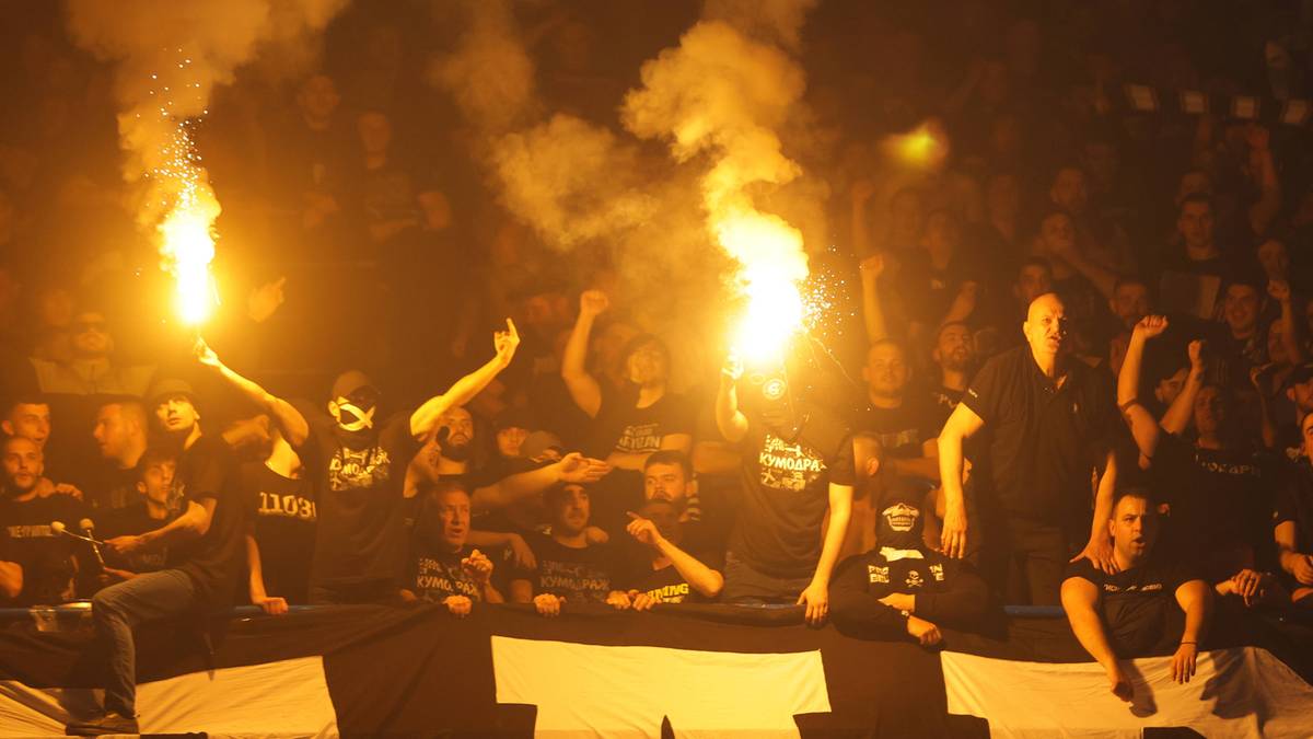 Tłum kibic&oacute;w na trybunach stadionu, z kt&oacute;rych wielu trzyma zapalone race i świece dymne, tworząc zadymioną atmosferę.