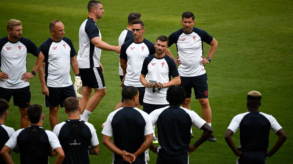 Grupa mężczyzn w strojach sportowych, prawdopodobnie piłkarzy i trener&oacute;w, stoi na zielonej murawie stadionu.