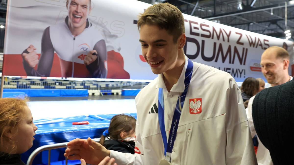 Młody mężczyzna w białej bluzie z kapturem i medalem na szyi podaje rękę widzowi. W tle widoczny jest baner z wizerunkiem sportowca i napisem "JESTEŚMY Z WAS DUMNI!". Wydarzenie sportowe.