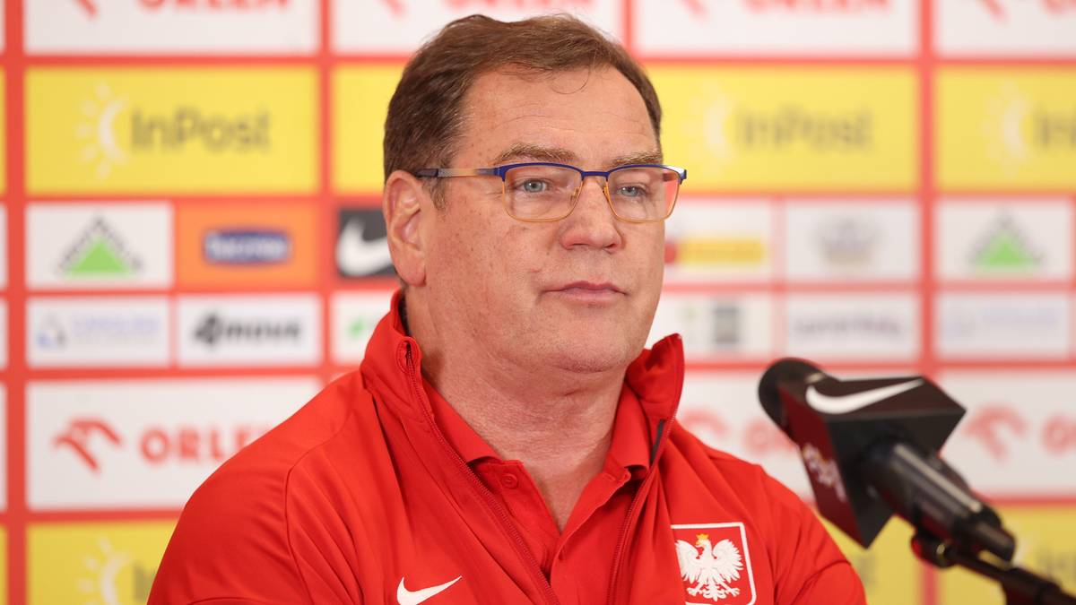 Jan Urban, w okularach, w czerwonej koszulce reprezentacji Polski, z herbem Polski, siedzi przed mikrofonem podczas konferencji prasowej.