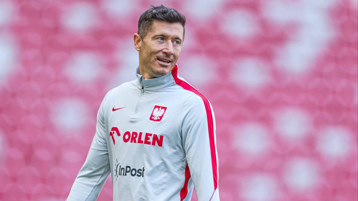 Piłkarz reprezentacji Polski w szarym stroju treningowym z czerwonymi elementami, patrzący w bok na tle czerwono-białych trybun stadionu.