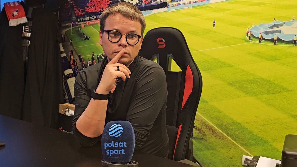 Mężczyzna w okularach siedzi przy biurku, opierając podbr&oacute;dek na dłoni, przed nim stoi mikrofon z logo Polsat Sport.