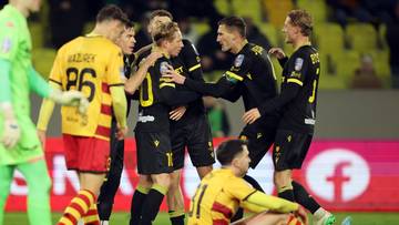 Piłkarze w czarnych strojach celebracji zdobytej bramki, jeden klęczy na boisku.