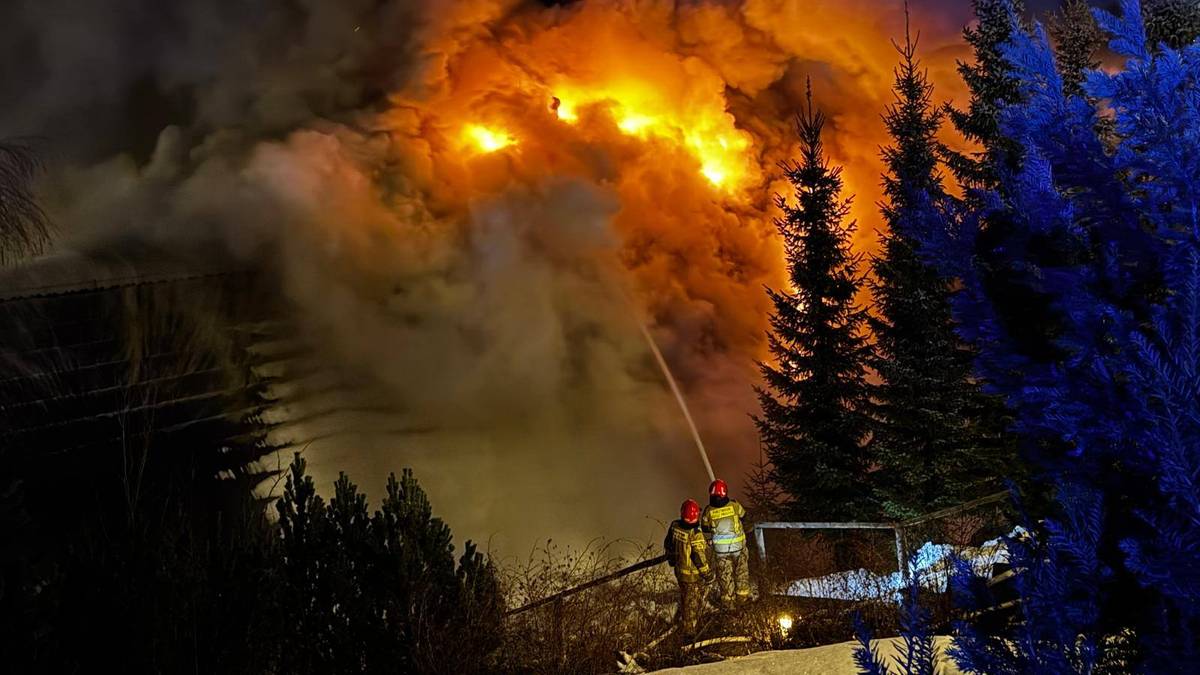 Dwaj strażacy walczą z płonącym budynkiem mieszkalnym w nocy, używając węża strażackiego. Widoczne są intensywne płomienie i gęsty dym.