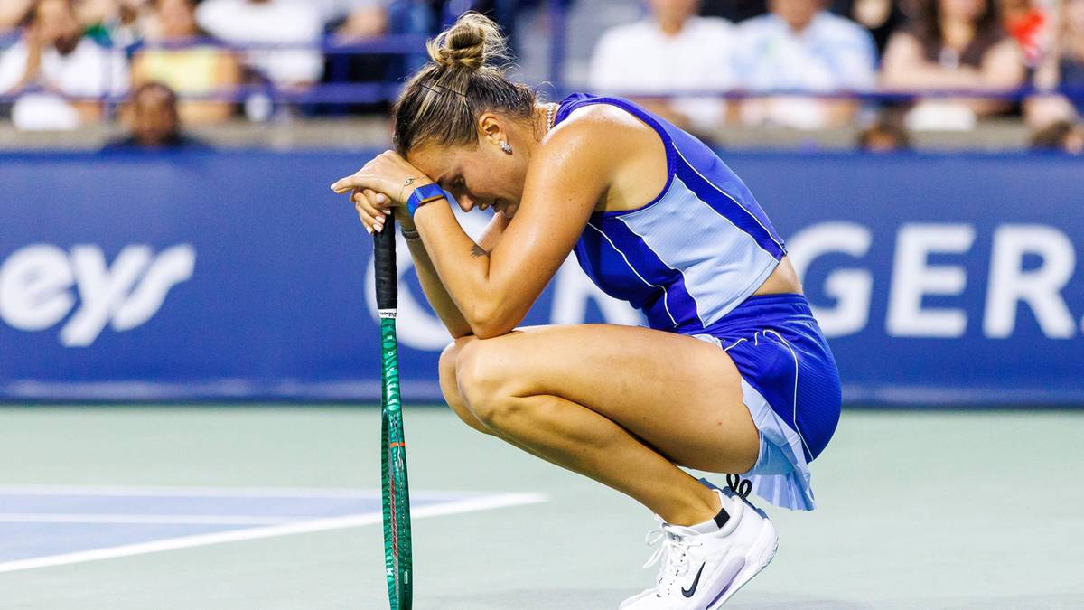 Zawodniczka tenisa w niebiesko-białym stroju sportowym kuca na korcie z głową opartą na rakiecie, wyrażając zmęczenie lub frustrację.