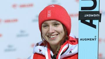 Uśmiechnięta skoczkini narciarska w czerwonej czapce i białej kurtce, trzymająca bukiet kwiat&oacute;w i dyplom.