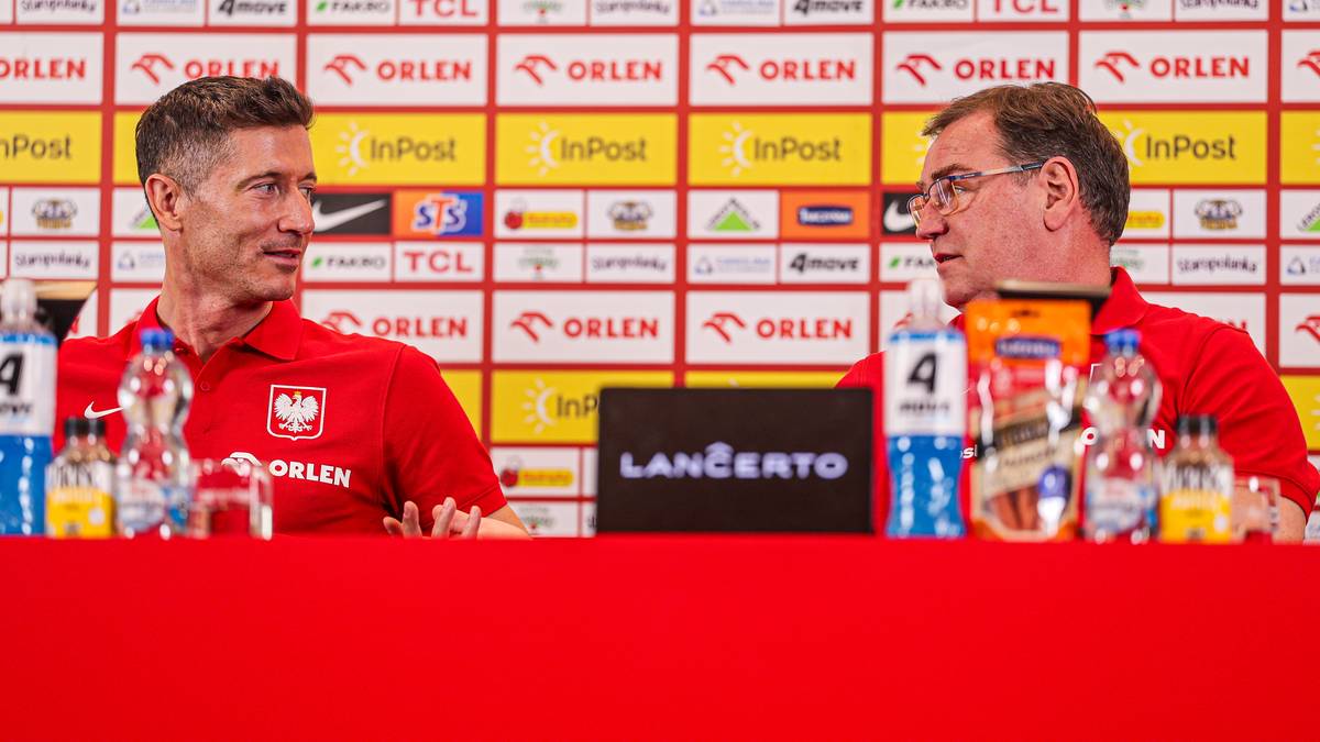 Dw&oacute;ch mężczyzn w czerwonych koszulkach z orłem na piersi siedzi przy stole. Na stole znajdują się butelki z wodą i napojami. W tle widoczna jest ściana z logo sponsor&oacute;w.