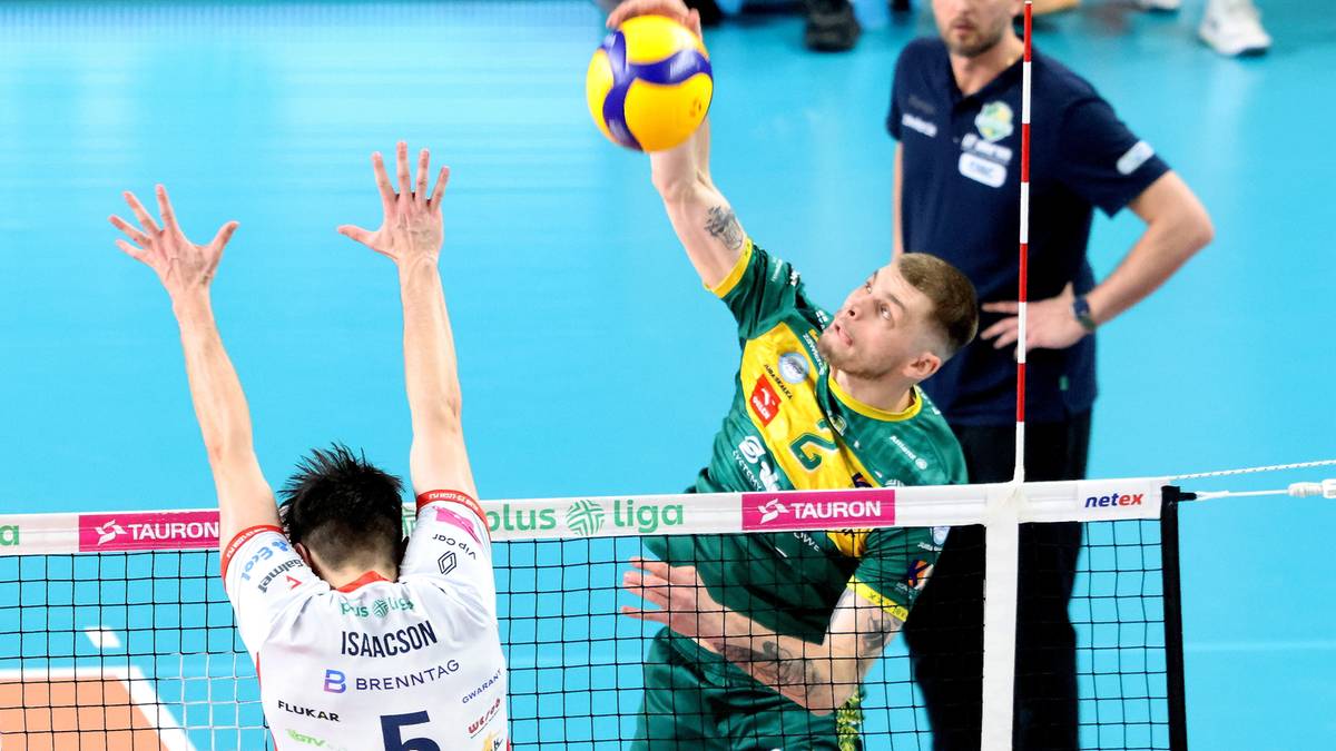 Siatkarz w zielonej koszulce wykonuje atak, a siatkarz w białej koszulce próbuje go blokować podczas meczu.