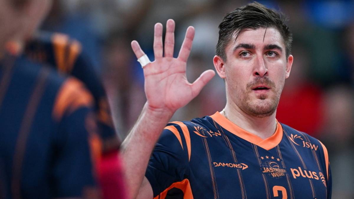 Siatkarz w niebiesko-pomarańczowej koszulce z numerem 2 unosi prawą dłoń.