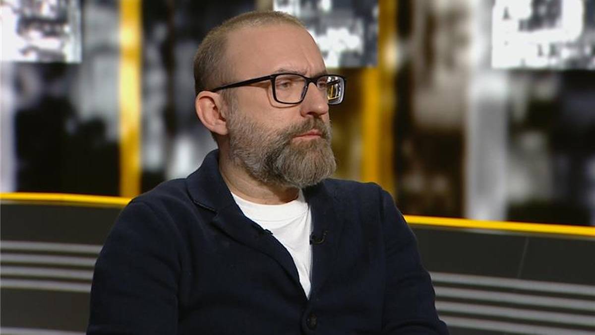 Mężczyzna z brodą i okularami siedzi na kanapie, ubrany w ciemną bluzę.