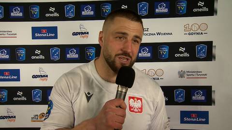 Piotr Zeszutek, mężczyzna z zarostem, przemawia do mikrofonu na tle ściany z logotypami.