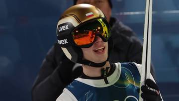 Skoczek narciarski w złotym kasku i goglach narciarskich, ubrany w biały kombinezon z numerem 30, trzyma narty.