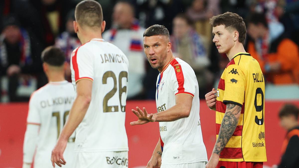 Trzech piłkarzy na boisku, dwóch w białych strojach, jeden w żółtym, rozmawiających podczas meczu.