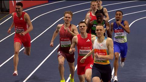 Sześciu lekkoatlet&oacute;w w czerwonych i białych strojach sportowych biegnie po niebieskim torze lekkoatletycznym. Dw&oacute;ch zawodnik&oacute;w na pierwszym planie, jeden w białej koszulce z czerwonymi i ż&oacute;łtymi pasami, drugi w czerwonej koszulce z numerem 34, wyprzedza pozostałych.