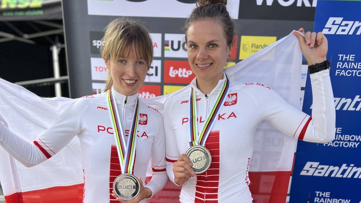 Dwie kobiety w strojach kolarskich z białymi i czerwonymi elementami, uśmiechają się do aparatu, każda z nich ma na szyi medal. Na strojach widnieje napis "POLSKA". Jedna z kobiet trzyma polską flagę.