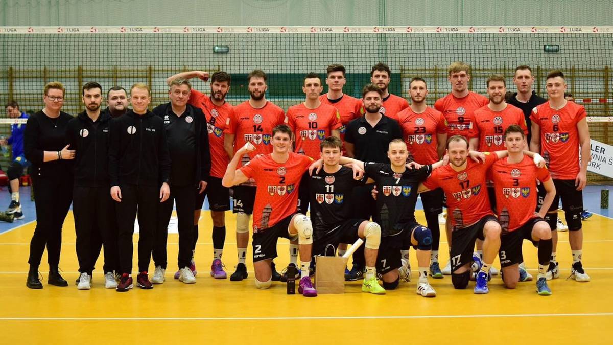 Drużyna siatkarska w strojach sportowych pozuje do zdjęcia na boisku siatkarskim.