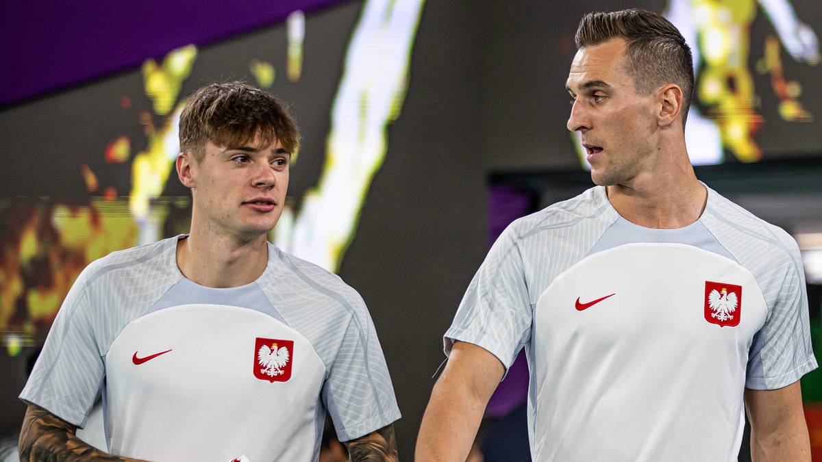 Dwóch piłkarzy reprezentacji Polski w szarych koszulkach treningowych z godłem Polski, patrzących w bok.