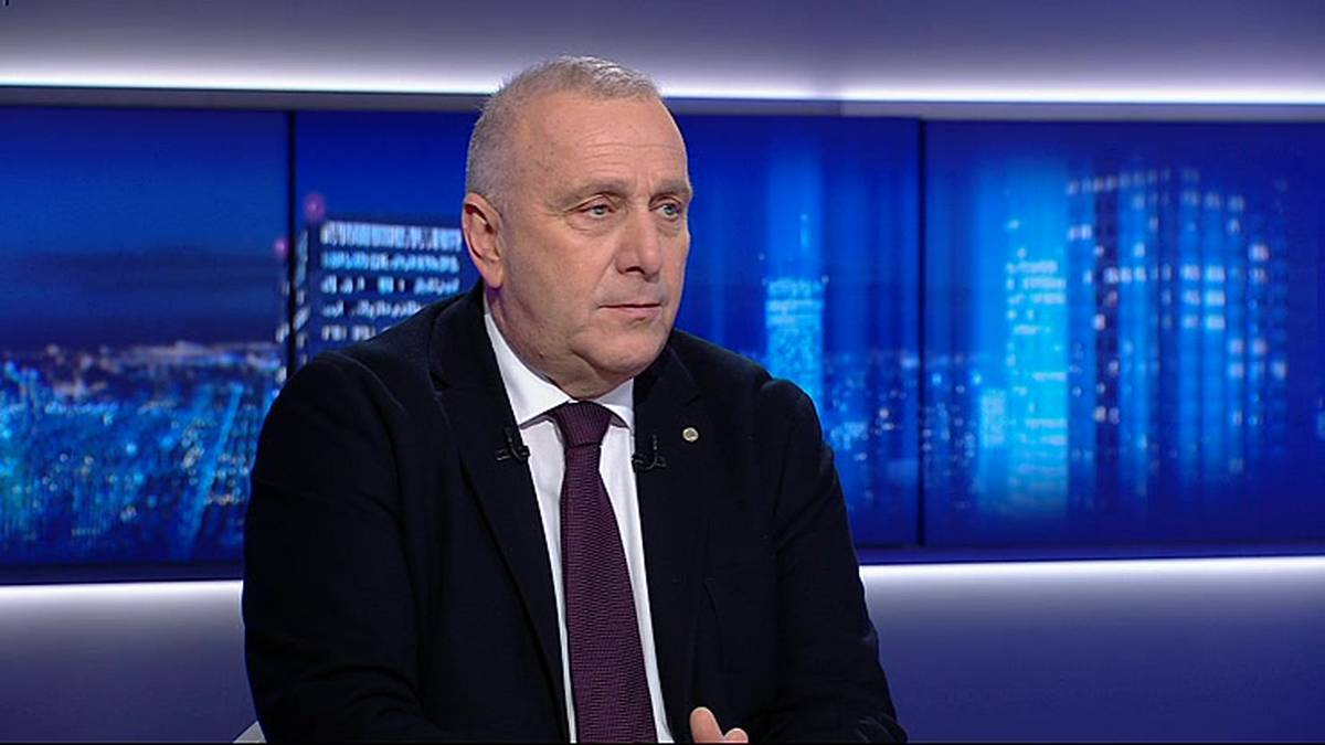 Mężczyzna w ciemnym garniturze i bordowym krawacie siedzi, patrząc lekko w bok. W tle za nim znajduje się duży ekran z rozmytym widokiem nocnej panoramy miasta z wieżowcami.